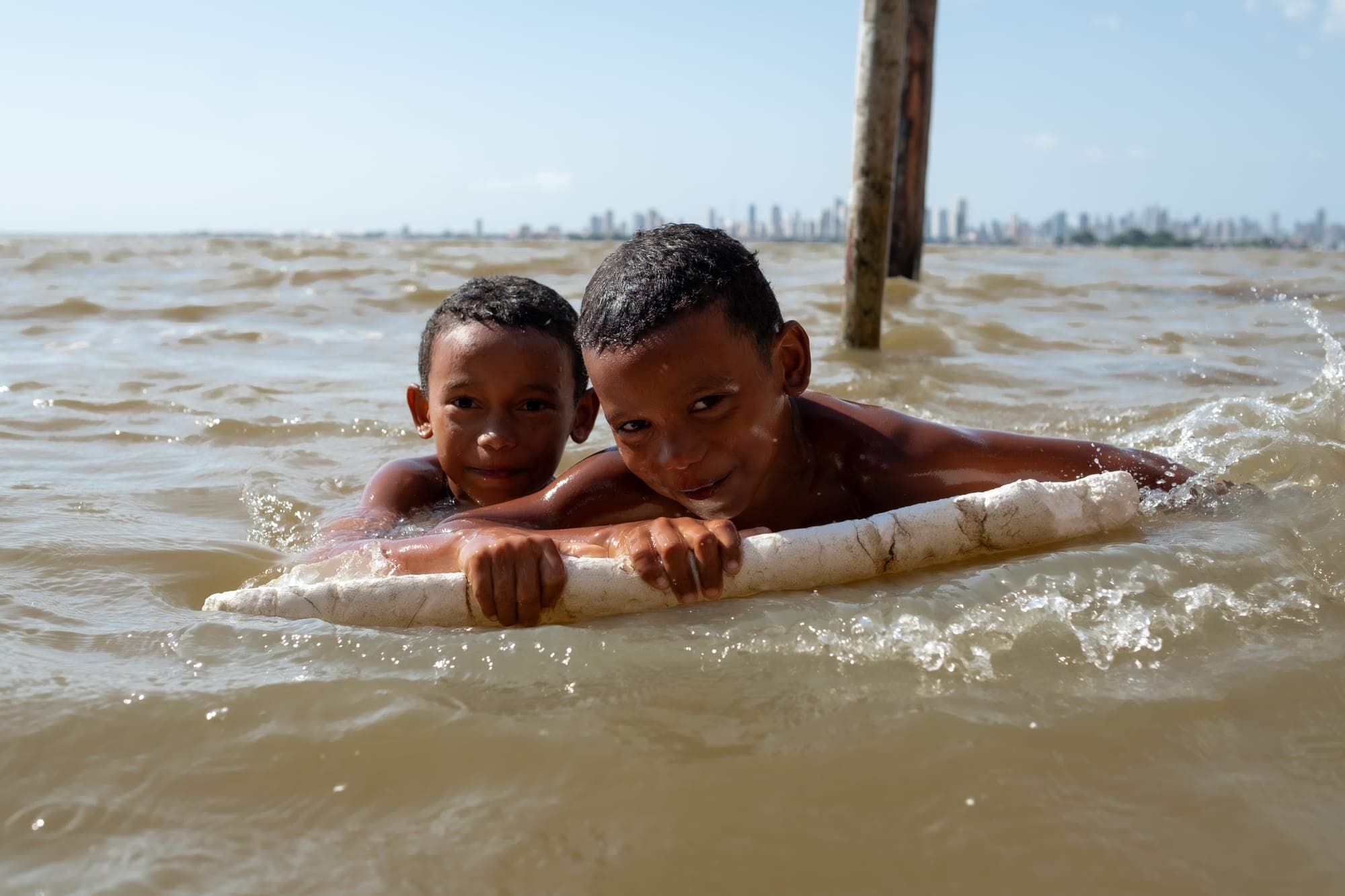 Dois meninos nadam sorrindo segurando um tronco no rio