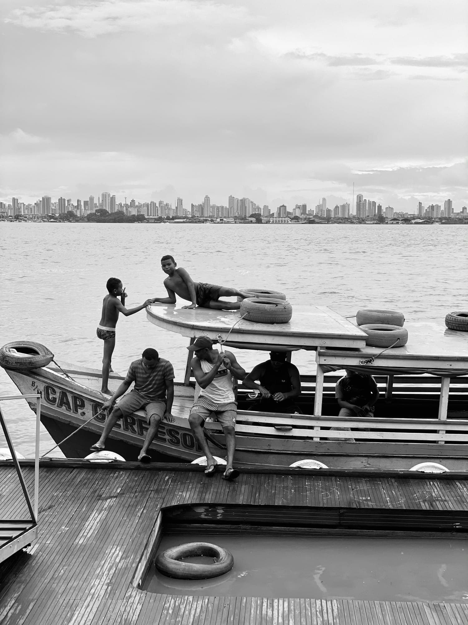 Meninos pulam de um barco atracado, com a cidade ao fundo, em preto e branco