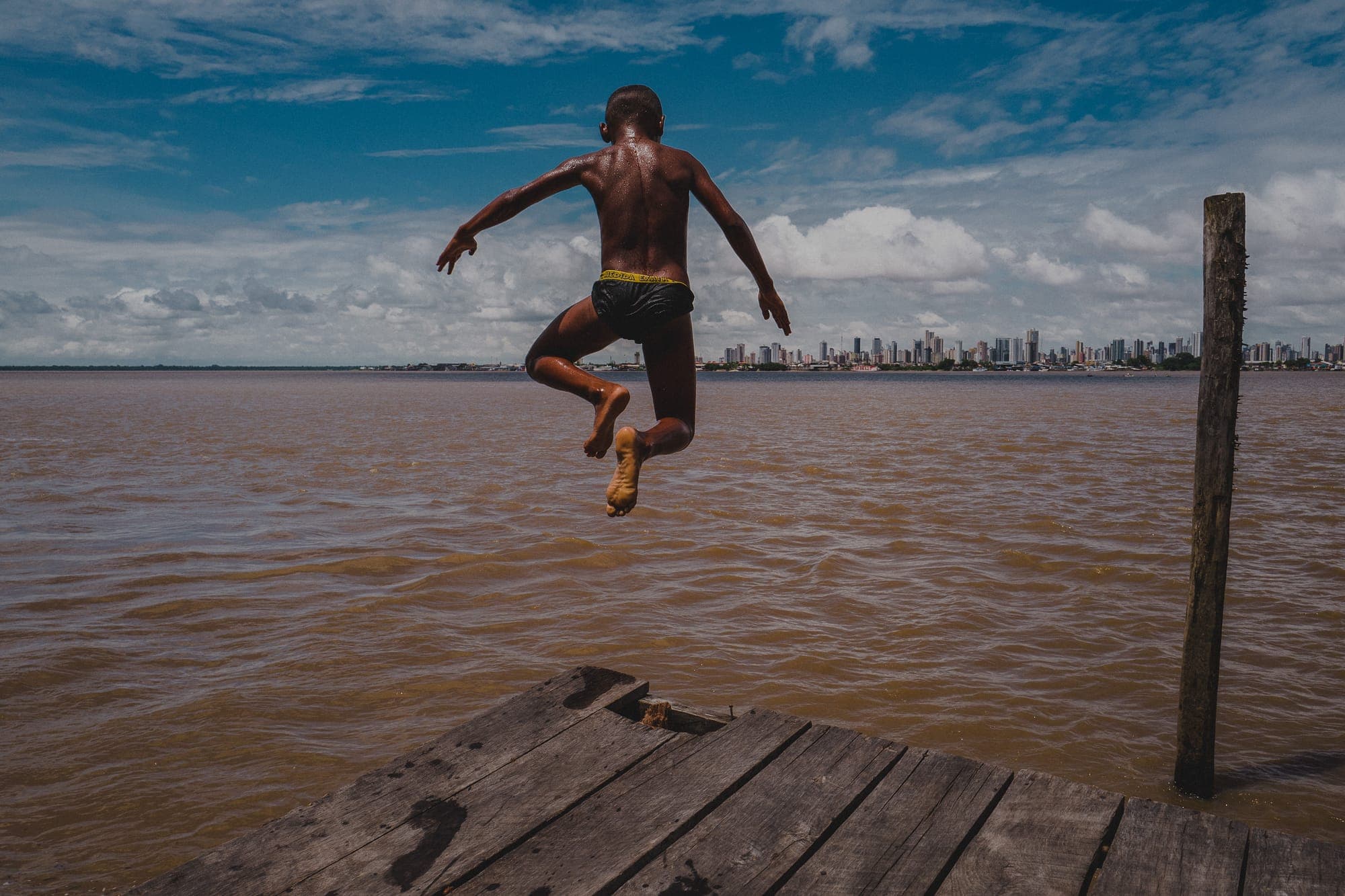 Menino salta de um trapiche para o rio, com a cidade ao fundo