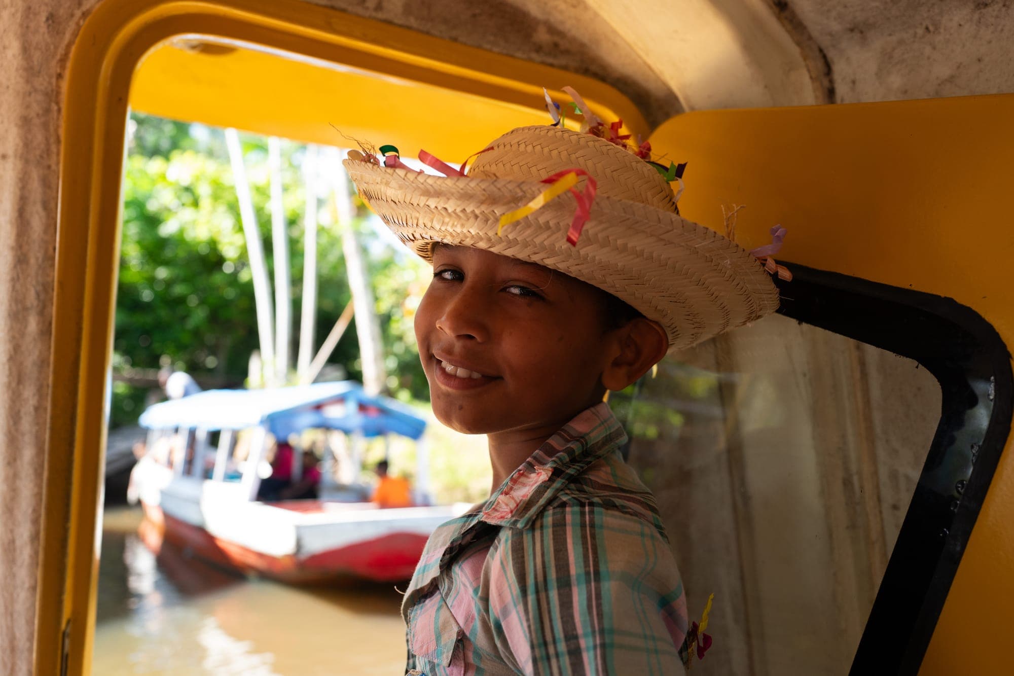 Menino de chapéu de palha sorri na janela de um barco