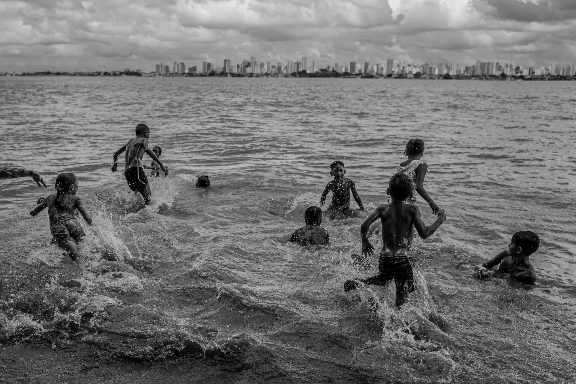 Crianças brincam na água do rio, com a cidade ao fundo, em preto e branco