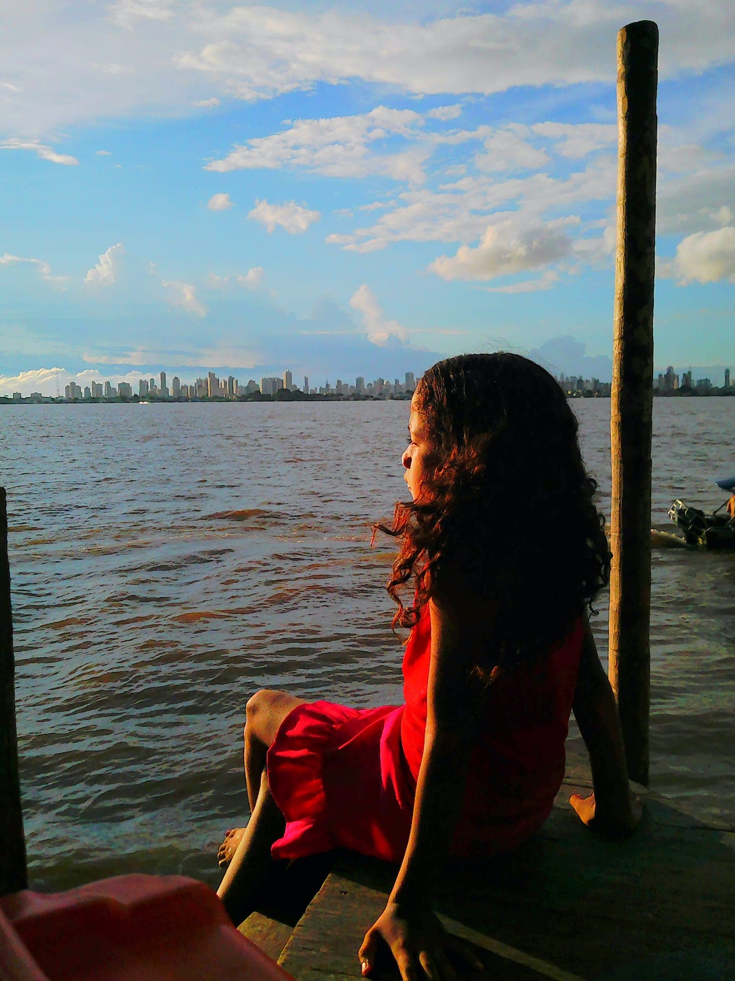 Menina sentada olhando o rio, com a cidade ao fundo