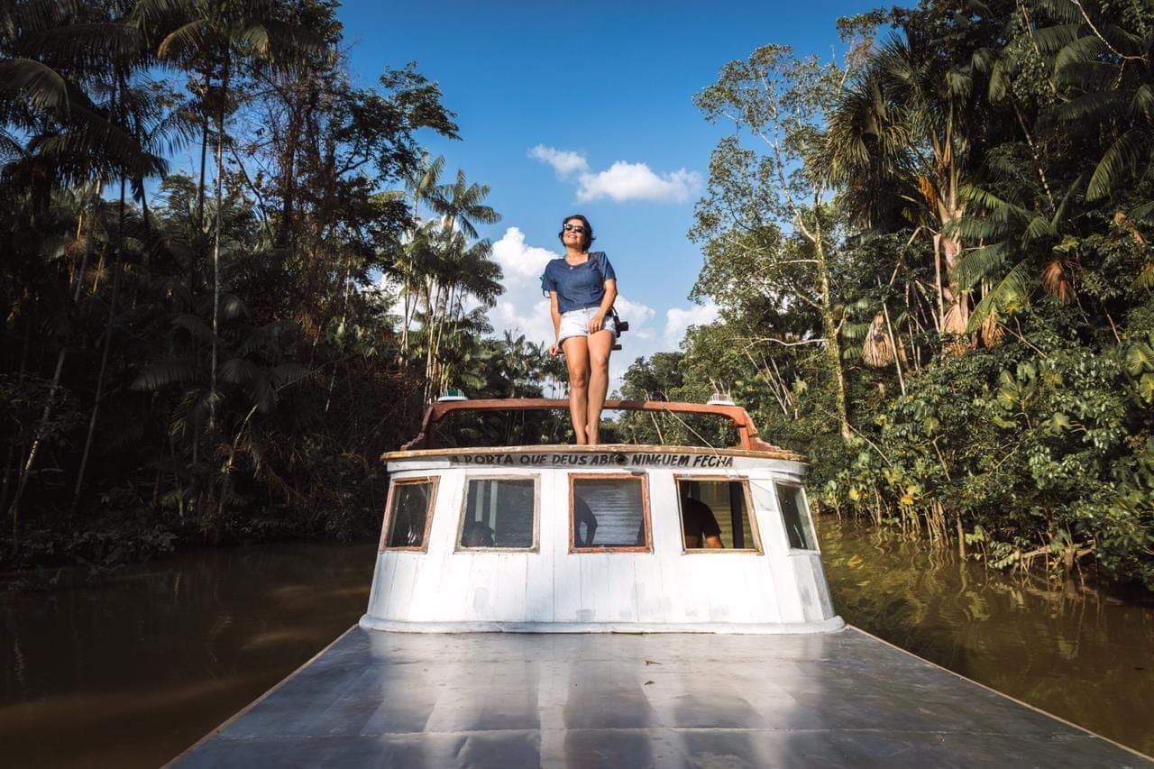 Mulher de pé no teto de um barco branco navegando por um igarapé