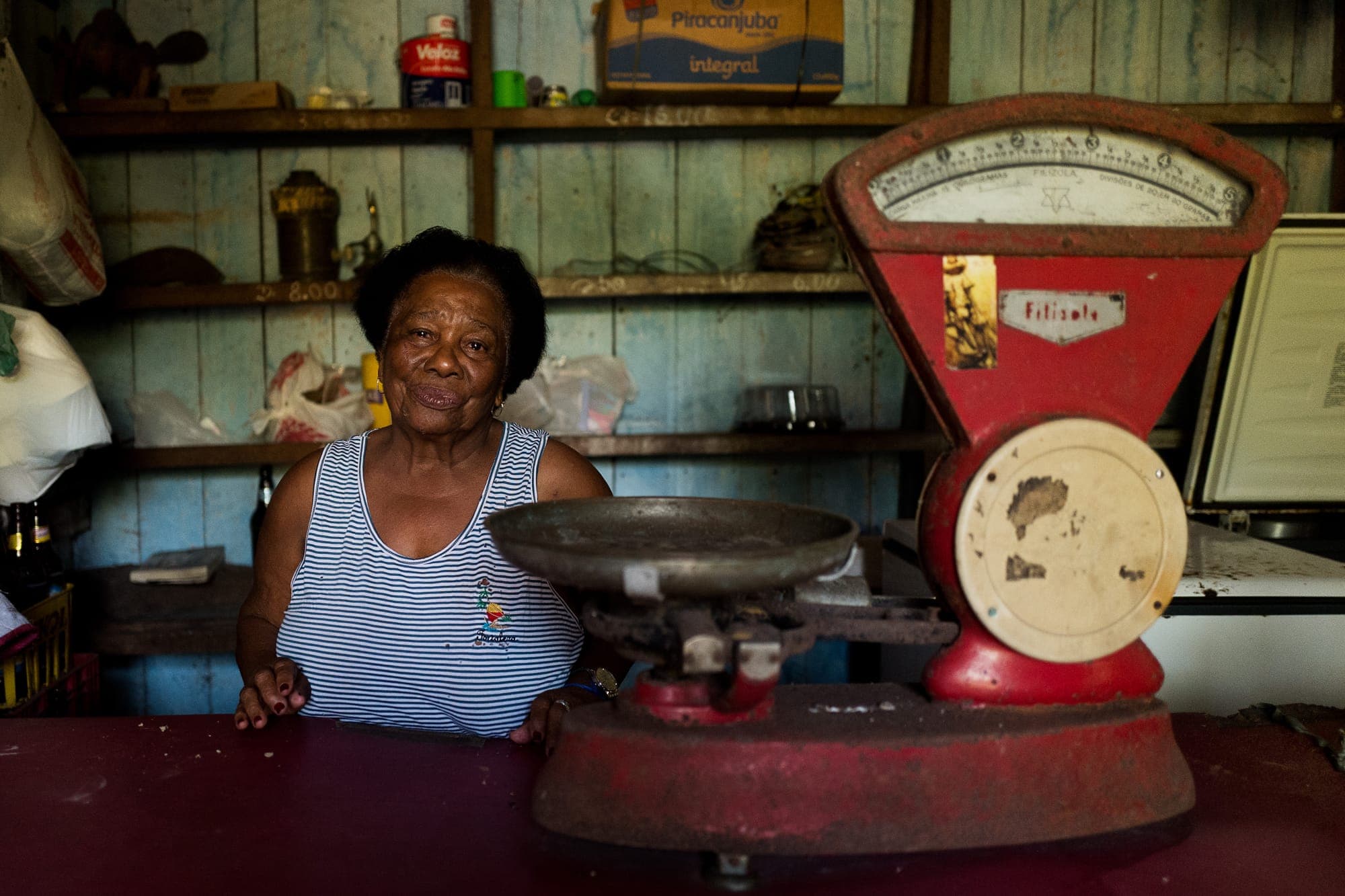 Moradora atrás do balcão de uma venda, ao lado de uma balança antiga