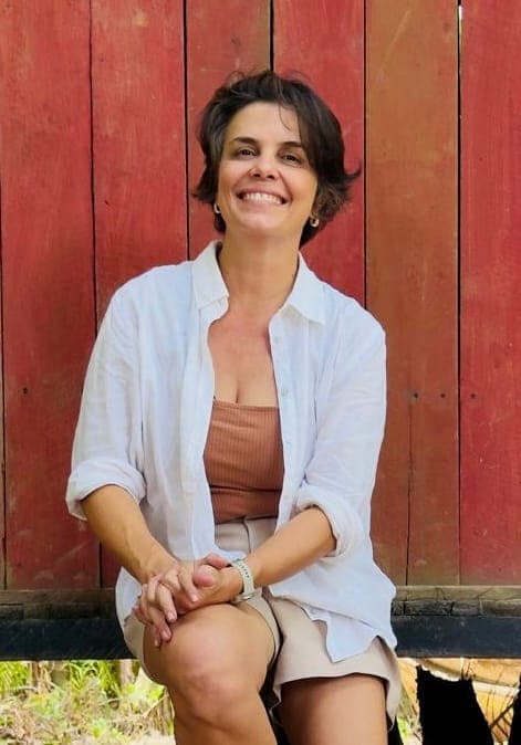 Grazi Caliman sorri, sentada diante de uma parede de madeira vermelha
